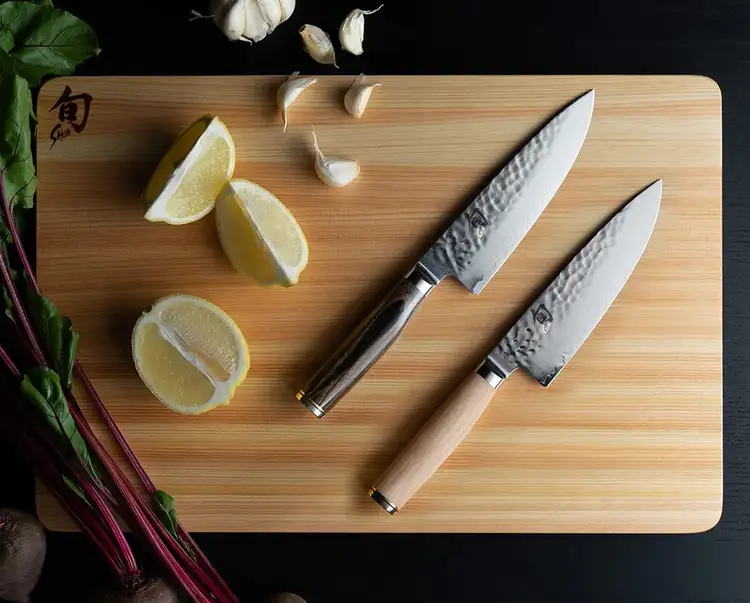 Two Premier 6" Chef's Knives, one with a blonde handle and the other with a walnut-stained handle, lie on a hinoki cutting board next to sliced lemons.