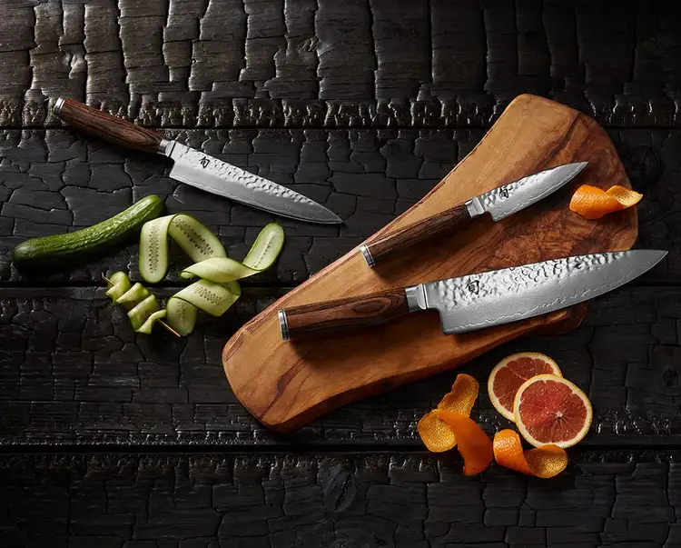 A group of three Premier knives lie on a serving board next to thinly sliced fruit and vegetables.