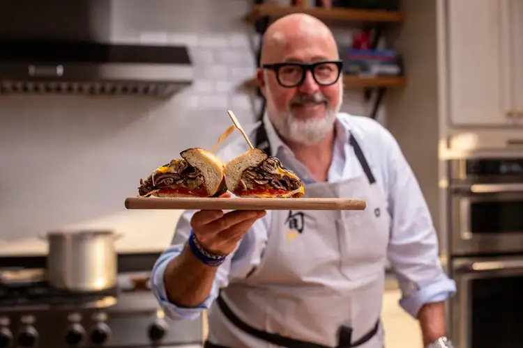 Chef Zimmern holds up his finished cheesesteak sandwich on a serving board.