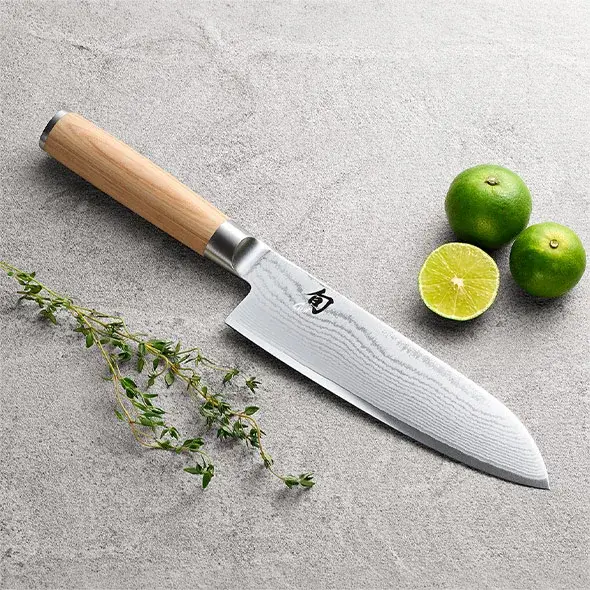 Santoku knife styled with herbs and limes on a stone surface.
