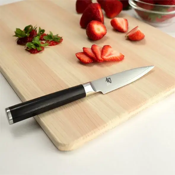 Paring knife beside sliced strawberries on a light wood board.