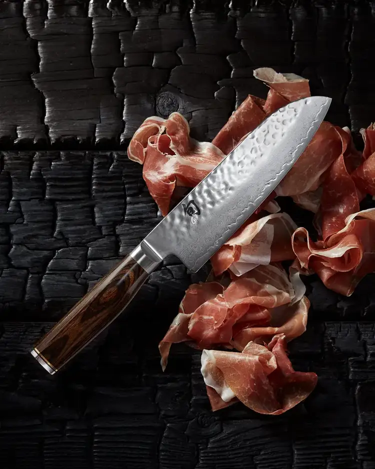 Shun Premier Santoku knife with prosciutto slices on a textured black wood background. Click to shop.