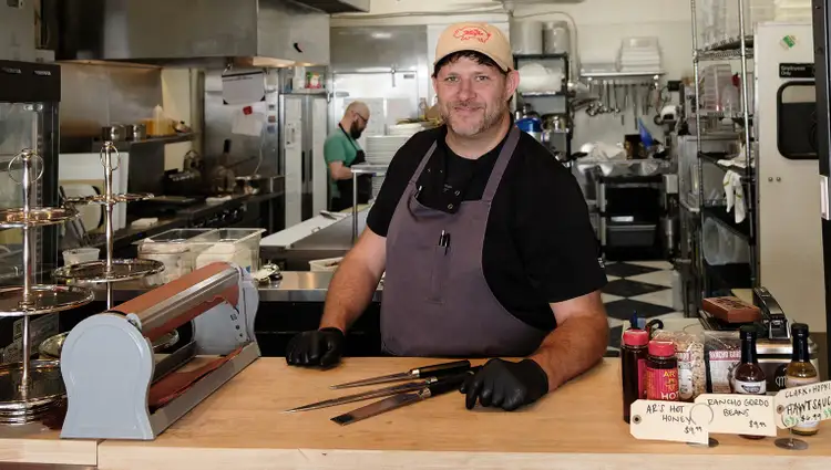 Chef Kyle Morse stands in The Mayor Butchery smiling at the camera, leaning on a wood countertop with three Shun knives in front of him.